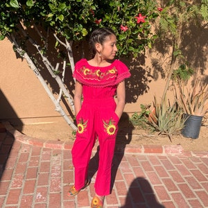 May include: A young person wearing a pink jumpsuit with embroidered sunflowers on the pockets and a floral embroidered neckline. The jumpsuit has a lace trim around the neckline. The person is wearing colorful sandals.