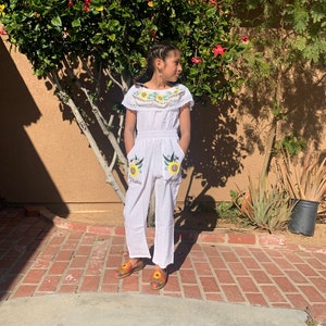 May include: A white jumpsuit with embroidered sunflowers and birds. The jumpsuit has a ruffled neckline and pockets. The model is wearing the jumpsuit with colorful sandals.