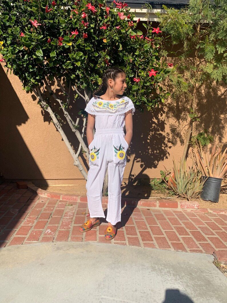 May include: A white jumpsuit with embroidered sunflowers and birds. The jumpsuit has a ruffled neckline and pockets. The model is wearing the jumpsuit with colorful sandals.
