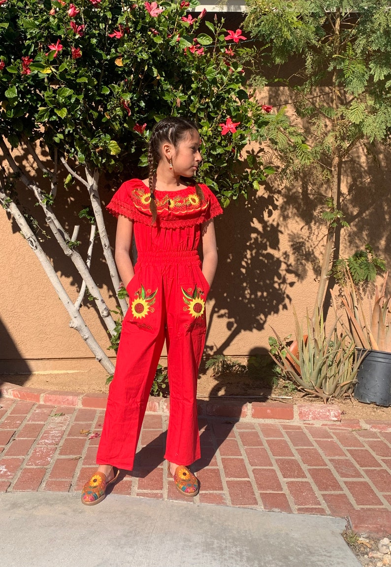 May include: A red jumpsuit with embroidered sunflowers and a ruffled neckline. The jumpsuit has short sleeves and is worn by a person with braided hair.