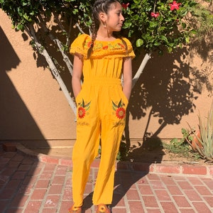 May include: A yellow jumpsuit with embroidered sunflowers and birds. The jumpsuit has a ruffled neckline and short sleeves. The model is wearing the jumpsuit with brown shoes.