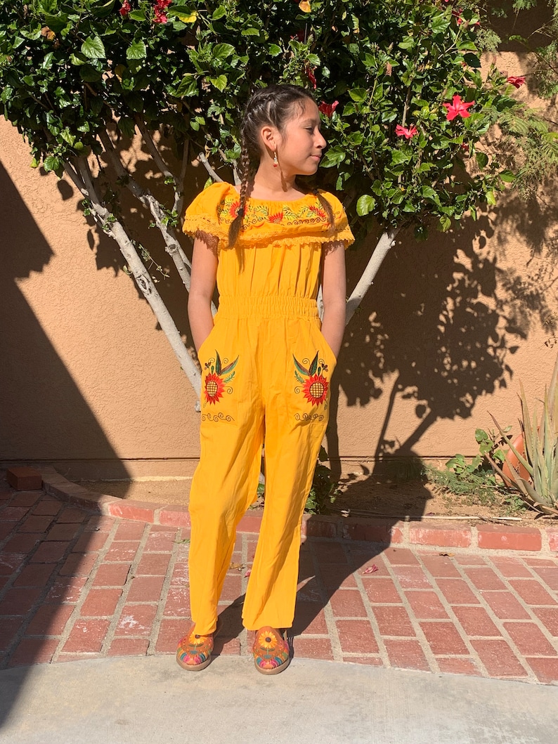 May include: A yellow jumpsuit with embroidered sunflowers and birds. The jumpsuit has a ruffled neckline and short sleeves. The model is wearing the jumpsuit with brown shoes.