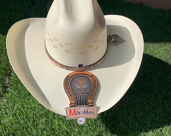 May include: A white straw cowboy hat with a brown leather band and a decorative horseshoe design. The hat has a label that says "MoksMan. Western Hats" and "XL".