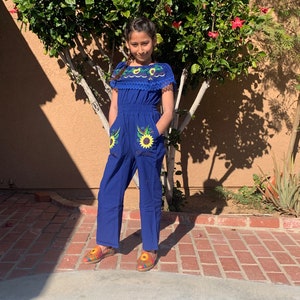 May include: A young person wearing a blue jumpsuit with embroidered sunflowers. The jumpsuit has a white lace trim around the neckline and short sleeves. The person is wearing colorful embroidered shoes.