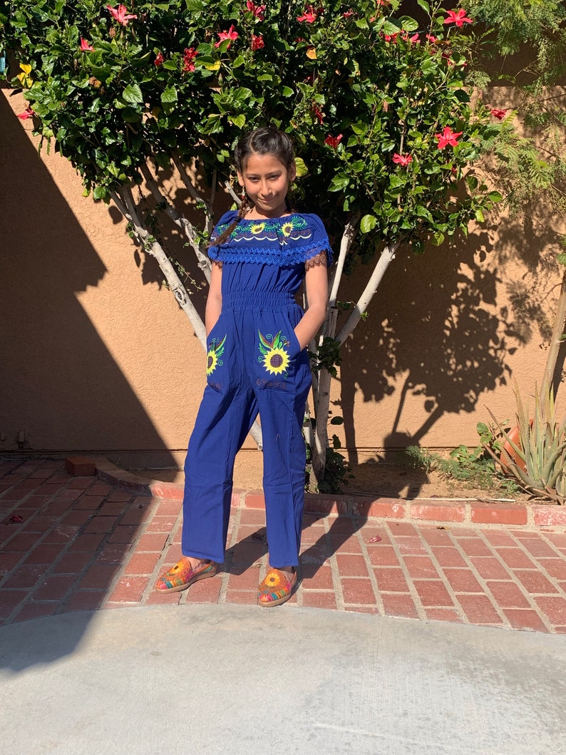 May include: A young person wearing a blue jumpsuit with embroidered sunflowers. The jumpsuit has a white lace trim around the neckline and short sleeves. The person is wearing colorful embroidered shoes.