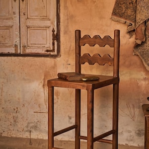 Könnte beinhalten: Ein hoher Barhocker aus Holz mit vier geraden Beinen, einer quadratischen Sitzfläche und einer Rückenlehne mit zwei horizontalen Leisten, die ein gewelltes, bogenförmiges Design aufweisen. Ein kleines, abgenutztes braunes Buch und ein metallischer Gegenstand liegen auf der Sitzfläche.