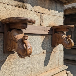 May include: Two wooden elephant head shelves fixed to a stone wall. The shelves are carved with detailed elephant features. The shelves are a warm brown colour, and are mounted on a stone wall.