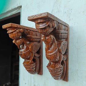 Peut inclure: Deux consoles en bois finement sculptées, teintées d'un brun chaud. Chacune présente des sculptures en relief détaillées d'oiseaux stylisés et de motifs floraux, parfaites pour ajouter une touche décorative à tout espace. Les consoles sont montées sur un mur clair.