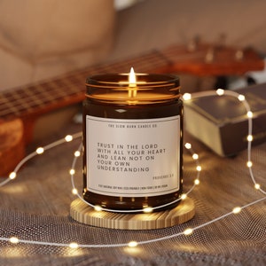 May include: A brown glass candle with a white label that reads "THE SLOW BURN CANDLE CO." and "TRUST IN THE LORD WITH ALL YOUR HEART AND LEAN NOT ON YOUR OWN UNDERSTANDING PROVERBS 3:5". The candle is lit and sitting on a wooden coaster.