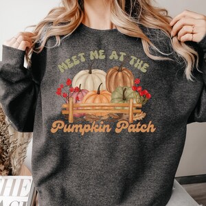 May include: A dark gray sweatshirt with the text "Meet me at the Pumpkin Patch" in white and orange lettering. The design features a small wooden fence with four pumpkins in various shades of orange, green, and white.