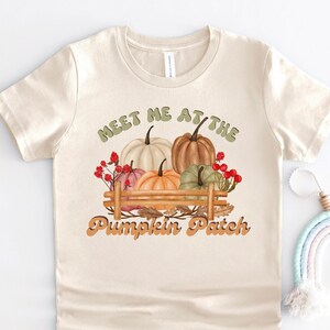 Puede incluir: Una camiseta beige con un gráfico de calabazas en una caja de madera. El texto "Meet me at the Pumpkin Patch" está impreso en una fuente retro.