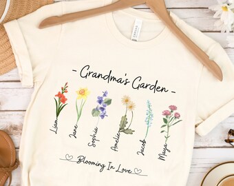 Può includere: Una maglietta bianca con il testo "Grandma's Garden" e un design floreale. I fiori sono etichettati con i nomi Liam, Jane, Sophia, Amelie, Jacob e Maya. Il testo "Blooming in Love" è sotto i fiori.