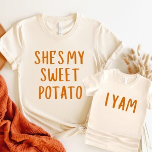 Op de afbeelding: Twee beige T-shirts met oranje tekst. Het grotere shirt zegt "She's my sweet potato" en het kleinere shirt zegt "I yam".