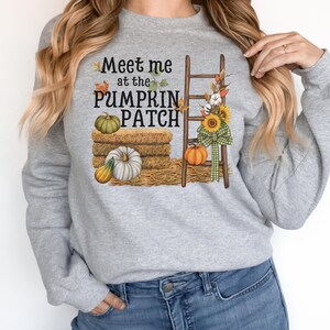 Könnte beinhalten: Grauer Pullover mit dem Text "Meet me at the PUMPKIN PATCH" und einer Grafik mit Kürbissen, Heuballen, einer Leiter und Sonnenblumen.
