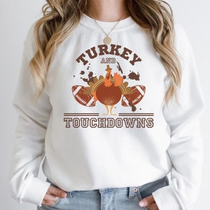 Könnte beinhalten: Weißer Pullover mit einem braunen Truthahn-Motiv und dem Text "TURKEY AND TOUCHDOWNS". Der Truthahn trägt einen Pilgerhut und hält zwei Fußbälle.