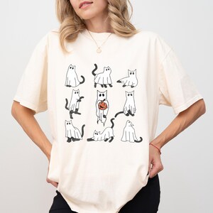 Puede incluir: Una camiseta blanca con un gr&aacute;fico de nueve gatos fantasma negros y blancos. Un gato sostiene una peque&ntilde;a calabaza naranja.