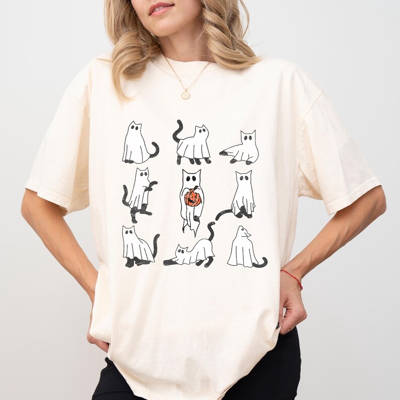 Puede incluir: Una camiseta blanca con un gr&aacute;fico de nueve gatos fantasma negros y blancos. Un gato sostiene una peque&ntilde;a calabaza naranja.