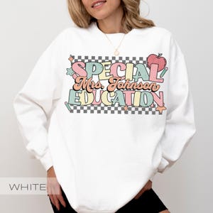 Op de afbeelding: Witte sweatshirt met de tekst "SPECIAL Mrs. Johnson EDUCATION" in pastelkleuren. Het ontwerp bevat een appel, sterren en een dambordpatroon. Een comfortabele top voor leraren.