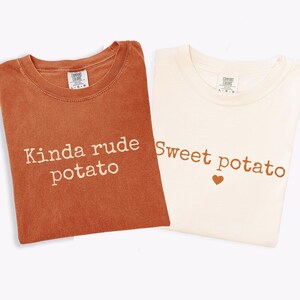 Könnte beinhalten: Zwei T-Shirts, eines in Rostfarbe und eines in Creme, werden gezeigt. Das rostfarbene Shirt trägt den weißen Schriftzug "Kinda rude potato". Das cremefarbene Shirt zeigt "Sweet potato" mit einem braunen Herzsymbol.