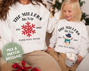 Könnte beinhalten: Passende weiße Sweatshirts mit einem Weihnachtsmotiv. Der Erwachsenen-Sweatshirt hat eine rote Schneeflocke und den Text "THE MILLERS Ski Trip EST. 2025 PARK CITY, UTAH". Der Kinder-Sweatshirt hat einen Schneemann und den Text "THE MILLERS Ski Trip EST. 2025 PARK CITY, UTAH".