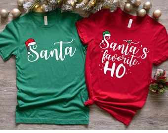 Può includere: Due magliette a tema natalizio. Una maglietta verde con un cappello di Babbo Natale rosso e la parola "Santa" in bianco. Una maglietta rossa con un cappello da elfo verde e il testo "Santa's favorite HO" in bianco.