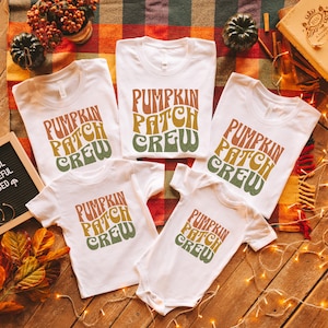 Pode incluir: Cinco camisetas brancas com o texto "Pumpkin Patch Crew" em uma fonte retrô com cores laranja, amarelo e verde. As camisetas estão dispostas em uma superfície de madeira marrom com uma manta xadrez e luzes de corda.