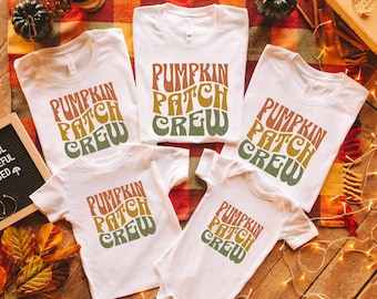 May include: Five white t-shirts with the text "Pumpkin Patch Crew" in a retro font with orange, yellow, and green colors. The shirts are arranged on a brown wooden surface with a plaid blanket and string lights.