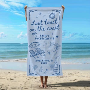 May include: A white beach towel with blue text that reads "Last toast on the coast Katie's Bachelorette Charleston, SC 2025". The towel features a beach umbrella, two lounge chairs, champagne glasses, and a bottle of champagne.