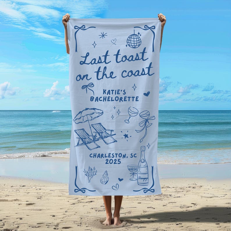 May include: A white beach towel with blue text that reads "Last toast on the coast Katie's Bachelorette Charleston, SC 2025". The towel features a beach umbrella, two lounge chairs, champagne glasses, and a bottle of champagne.