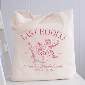 Op de afbeelding: Een witte canvas tote bag met een roze grafisch ontwerp. De grafiek toont een vrouw in een cowboyhoed die in een martini-glas zit met een discobal en een ukelele. De tekst luidt "Last Rodeo Est. 2025 Nicole's Bachelorette Nashville, TN".