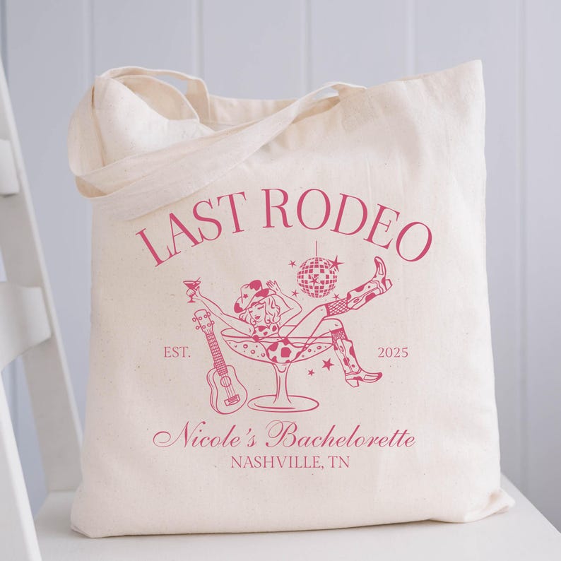 Op de afbeelding: Een witte canvas tote bag met een roze grafisch ontwerp. De grafiek toont een vrouw in een cowboyhoed die in een martini-glas zit met een discobal en een ukelele. De tekst luidt "Last Rodeo Est. 2025 Nicole's Bachelorette Nashville, TN".