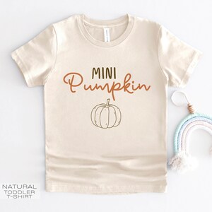 Pu&ograve; includere: Una maglietta beige per bambini piccoli con il testo "MINI Pumpkin" in lettere marroni e un semplice disegno di una zucca.