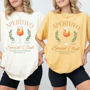 May include: Two women wearing white and yellow t-shirts with the text "Aperitivo Social Club" and a graphic of a cocktail glass with an orange drink. The text also includes "Est. 2024" and "Taylor's Bachelorette Savannah, GA".