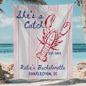 May include: Beach towel with a pink and white striped background. The towel features a red lobster illustration inside a dotted oval, with the text "She's a Catch" and "Katie's Bachelorette Charleston, SC".
