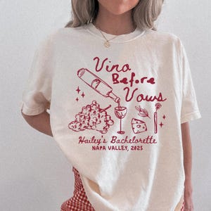 以下が含まれることがあります： クリーム色のTシャツ。バーガンディ色のグラフィックで「Vina Before Vows」とワインをテーマにしたイラストが描かれています。「Hailey's Bachelorette, Napa Valley, 2025」の文字もプリントされています。