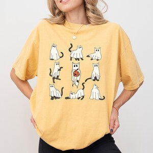 Puede incluir: Una camiseta amarilla con un estampado gr&aacute;fico de nueve gatos blancos vestidos de fantasmas. Los gatos est&aacute;n en varias poses, incluyendo sosteniendo una calabaza.