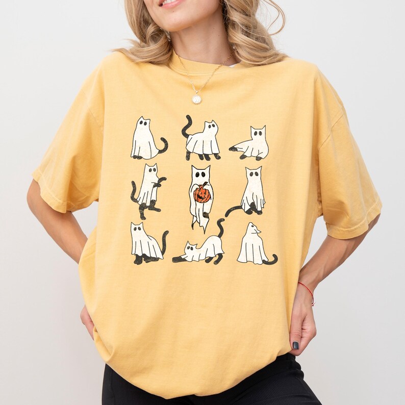 Puede incluir: Una camiseta amarilla con un estampado gr&aacute;fico de nueve gatos blancos vestidos de fantasmas. Los gatos est&aacute;n en varias poses, incluyendo sosteniendo una calabaza.