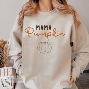 Pu&ograve; includere: Una felpa beige con il testo "Mama Pumpkin" in lettere marroni e arancioni. Un contorno marrone di una zucca si trova sotto il testo.