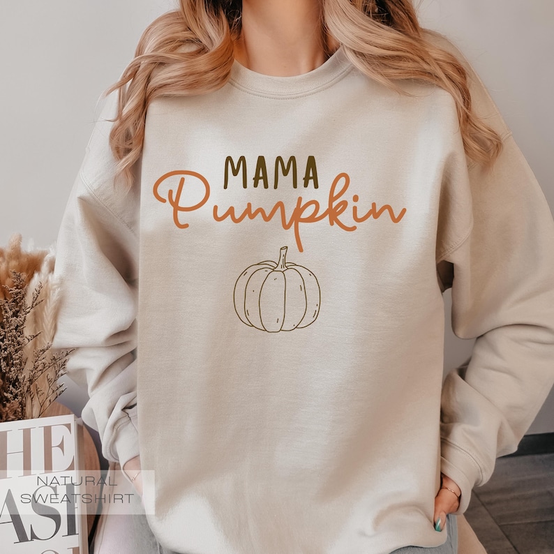 Pu&ograve; includere: Una felpa beige con il testo "Mama Pumpkin" in lettere marroni e arancioni. Un contorno marrone di una zucca si trova sotto il testo.