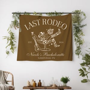 Pu&ograve; includere: Un arazzo marrone con il testo "Last Rodeo" in bianco. L'arazzo presenta una silhouette bianca di una donna con un cappello da cowboy seduta in un bicchiere da martini con una palla da discoteca e un ukulele. Il testo "Est. 2024" e "Nicole's Bachelorette Nashville, TN" &egrave; anche in bianco.