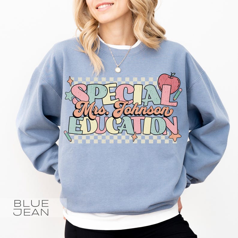 Op de afbeelding: Een lichtblauwe sweatshirt met de tekst "SPECIAL Mrs. Johnson EDUCATION" in een retro lettertype. Het ontwerp bevat een appel, sterren en potloden. De woorden "BLUE JEAN" staan onderaan gedrukt.