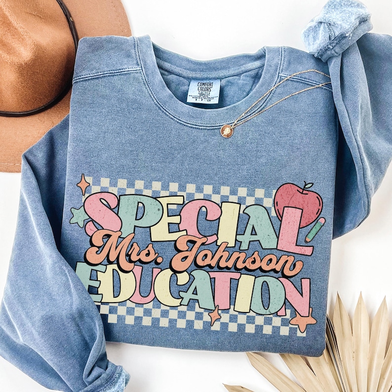 Op de afbeelding: Een blauwe crewneck sweatshirt met de tekst "SPECIAL Mrs. Johnson EDUCATION" in een kleurrijk retro lettertype. Het ontwerp bevat een appel en een potlood. De sweatshirt heeft een vintage, gewassen look.