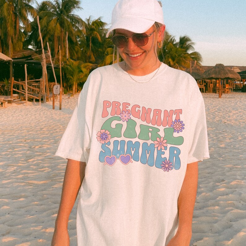 Puede incluir: Camiseta blanca con un gr&aacute;fico de estilo retro que dice "Pregnant Girl Summer" en letras rosas, verdes y azules con flores y corazones.