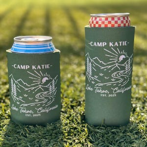 May include: Two green can coolers with a white mountain scene and the text "Camp Katie - Lake Tahoe, California EST. 2025".