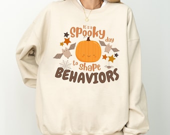 May include: A cream-colored sweatshirt with a graphic print of a smiling pumpkin, bats, and stars. The text reads "It's a spooky day to shape behaviors".
