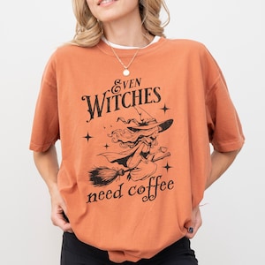May include: Orange t-shirt with a black graphic of a witch flying on a broomstick. The text "Even Witches need coffee" is printed on the shirt.