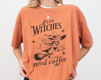 Puede incluir: Camiseta naranja con un gráfico negro de una bruja volando en una escoba. El texto "Even Witches need coffee" está impreso en la camiseta.