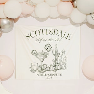 Puede incluir: Una pancarta crema con el texto "SCOTTSDALE Before the Veil" y "KATIE'S BACHELORETTE 2024". La pancarta presenta ilustraciones de una margarita, un cactus, una botella de tequila y una bola de discoteca. Globos blancos, plateados y rosas rodean la pancarta.
