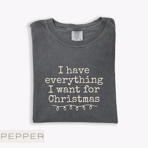 May include: A dark grey t-shirt with the white text "I have everything I want for Christmas". The shirt has a crew neck and a tag that says "Comfort Colors". A string of lights is below the text.