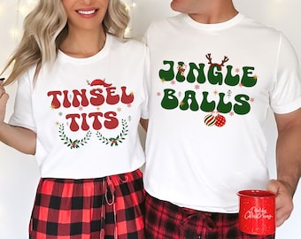 May include: A couple wearing matching white t-shirts with a Christmas-themed design. The woman's shirt says "Tinsel Tits" in red and green lettering with a Santa hat and snowflakes. The man's shirt says "Jingle Balls" in green and red lettering with a reindeer and ornaments.